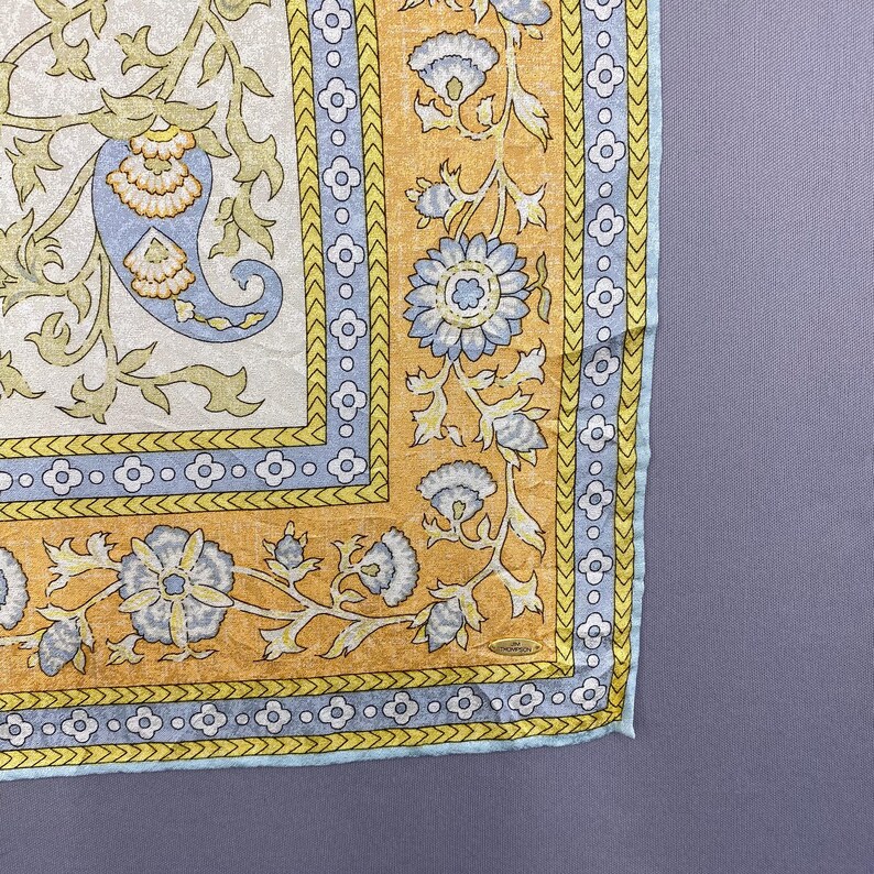 Puede incluir: Un pa&ntilde;uelo de seda cuadrado con un centro de color crema con un patr&oacute;n de cachemira azul y enredaderas verdes. El borde es de un amarillo mostaza vibrante con motivos florales azules y hojas verdes, enmarcado por una banda azul claro.