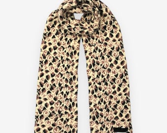 May include: A beige and brown patterned scarf. The scarf features a repeating pattern of brown and black shapes on a beige background. The scarf is made of a soft, knitted material and has a tag on one end.