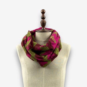 May include: A knitted infinity scarf in shades of fuchsia, olive green, and burgundy. The scarf features a geometric pattern and is displayed on a mannequin. The soft, warm scarf is a cozy accessory for cold weather.
