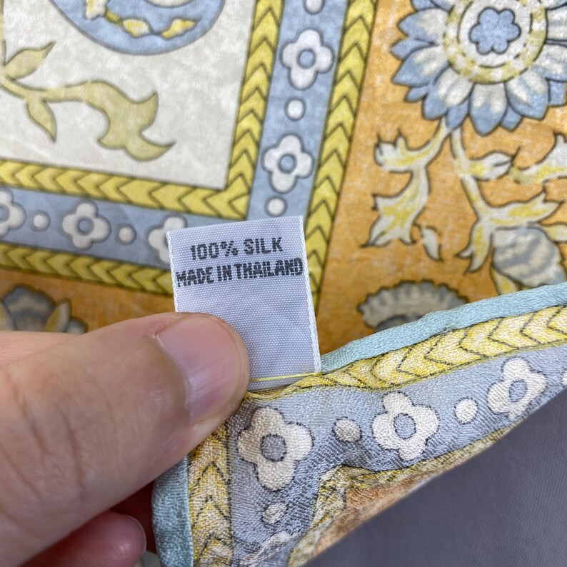 Puede incluir: Un pa&ntilde;uelo de seda con un estampado floral en tonos amarillo, azul claro y blanco. Una etiqueta de tela blanca est&aacute; unida al borde, impresa con el texto 100% SILK MADE IN THAILAND. El pa&ntilde;uelo tiene un borde decorativo con formas geom&eacute;tricas.
