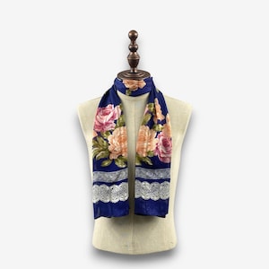 Puede incluir: Una bufanda azul marino con un estampado floral de rosas rosas y melocotón y hojas verdes. La bufanda tiene un borde de encaje blanco. La bufanda se exhibe en un maniquí.