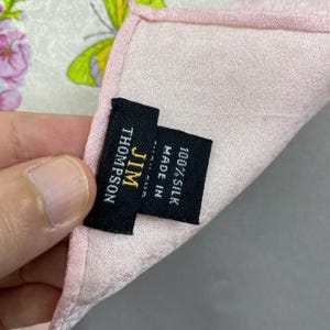 Puede incluir: Un pa&ntilde;uelo de seda rosa con un estampado floral y de mariposas. El pa&ntilde;uelo tiene una etiqueta que dice "JIM THOMPSON" y "100% SEDA MADE IN". Los bordes son de un rosa m&aacute;s oscuro.