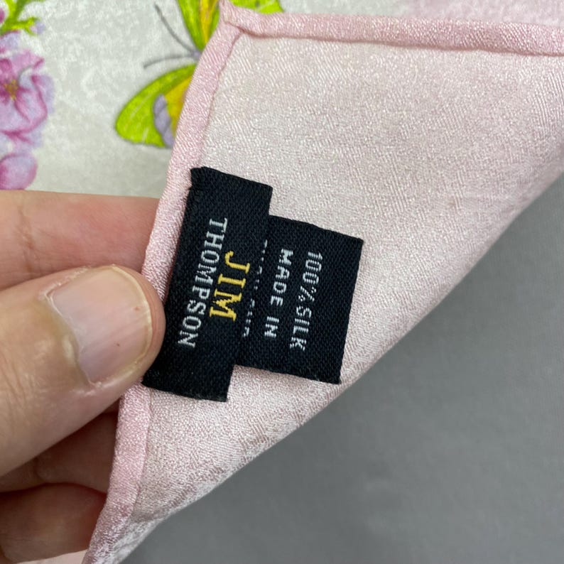 Puede incluir: Un pa&ntilde;uelo de seda rosa con un estampado floral y de mariposas. El pa&ntilde;uelo tiene una etiqueta que dice "JIM THOMPSON" y "100% SEDA MADE IN". Los bordes son de un rosa m&aacute;s oscuro.
