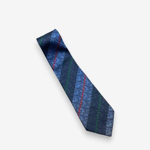 Könnte beinhalten: Eine blaue Krawatte mit Streifenmuster. Die Krawatte hat abwechselnde Streifen in Dunkelblau, Hellblau und ein Muster aus roten und grünen floralen Designs. Die Krawatte ist diagonal vor einem weißen Hintergrund abgebildet.
