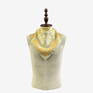 Puede incluir: Un pa&ntilde;uelo cuadrado con un patr&oacute;n floral en amarillo y blanco con detalles de l&iacute;neas grises. El pa&ntilde;uelo est&aacute; doblado en tri&aacute;ngulo y exhibido sobre un maniqu&iacute;, mostrando un borde decorativo con motivos florales repetidos y l&iacute;neas geom&eacute;tricas.
