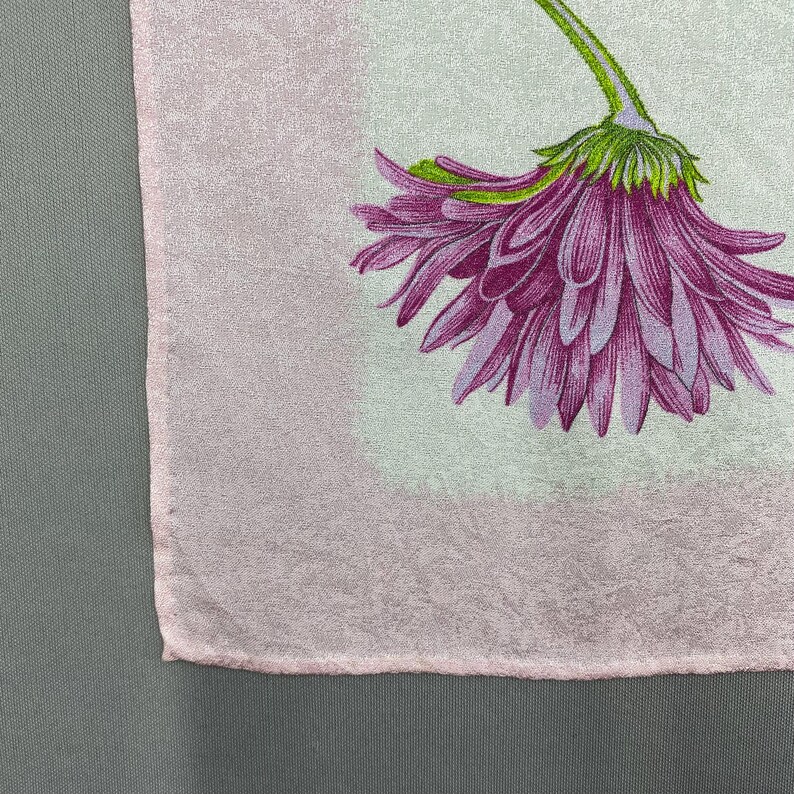 Puede incluir: Un textil rosa claro con un gran estampado floral morado detallado. La flor tiene un tallo y una base verdes, con p&eacute;talos en varios tonos de morado. El textil tiene un aspecto sutil y texturizado.