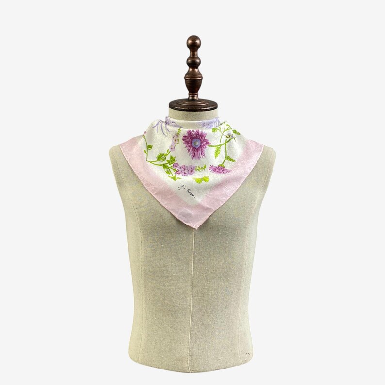 Puede incluir: Un pa&ntilde;uelo cuadrado con estampado floral y borde rosa se muestra sobre un maniqu&iacute;. El pa&ntilde;uelo presenta flores moradas y rosas con hojas verdes sobre un fondo blanco. La tela parece ligera.