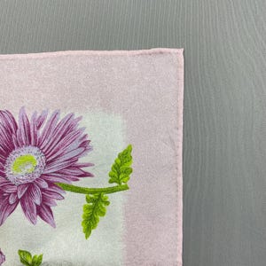 Puede incluir: Un pa&ntilde;uelo cuadrado de color rosa claro con un dise&ntilde;o floral. El dise&ntilde;o presenta una gran flor morada con un centro amarillo y hojas verdes. El pa&ntilde;uelo tiene un aspecto suave y texturizado.