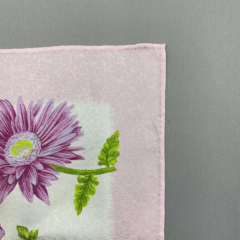 Puede incluir: Un pa&ntilde;uelo cuadrado de color rosa claro con un dise&ntilde;o floral. El dise&ntilde;o presenta una gran flor morada con un centro amarillo y hojas verdes. El pa&ntilde;uelo tiene un aspecto suave y texturizado.