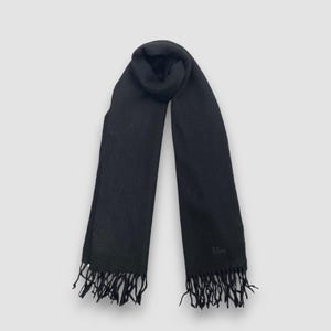 May include: A black scarf with fringe detailing. The scarf is made of a soft material and features the word "Dior" embroidered near the bottom. The scarf is a solid black color and is displayed against a white background.