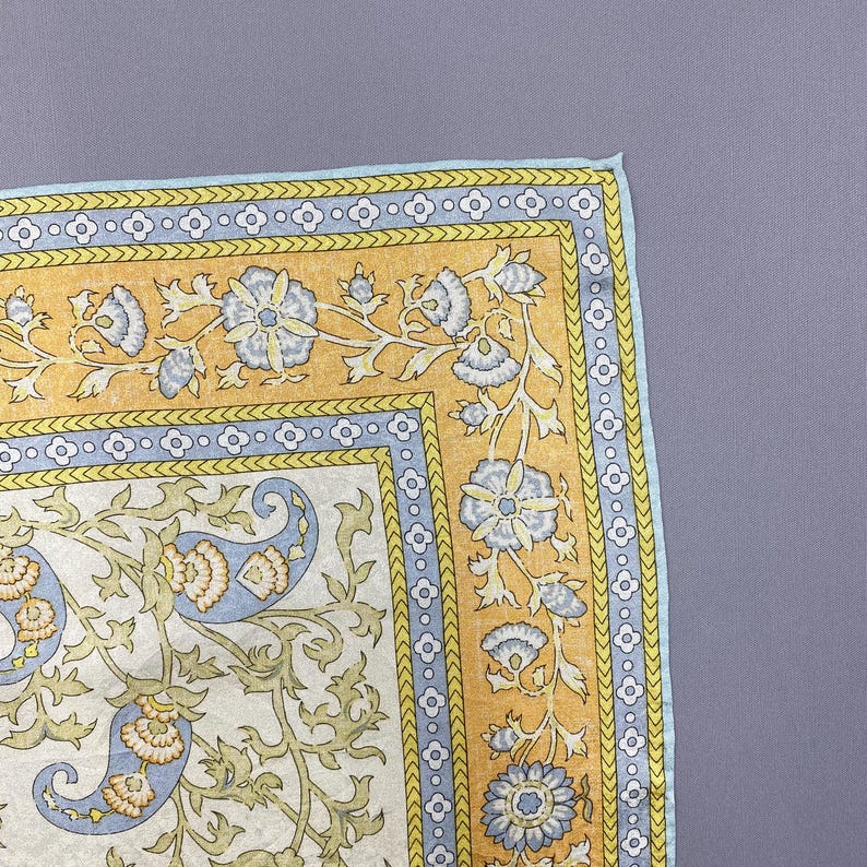 Puede incluir: Un pa&ntilde;uelo de bolsillo cuadrado de tela con un centro de color crema y un patr&oacute;n de cachemira en azul claro y verde oliva. El borde consta de una banda azul claro con motivos de flores blancas y un marco amarillo con enredaderas azules.