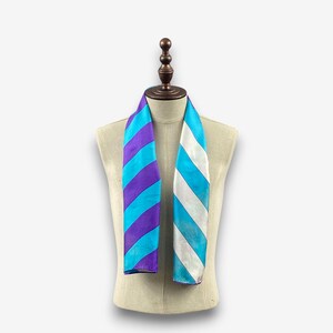 Puede incluir: Una bufanda colorida con rayas diagonales moradas, turquesas y celeste por un lado, y rayas turquesas y blancas por el otro. La bufanda se exhibe en un maniquí. La bufanda está hecha de un material sedoso.