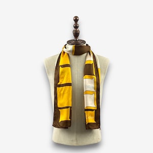 Puede incluir: Un pañuelo de seda con un estampado geométrico en amarillo, marrón y blanco. El pañuelo se exhibe en un maniquí. El pañuelo tiene un borde marrón y la palabra "Echo" está impresa en el pañuelo.