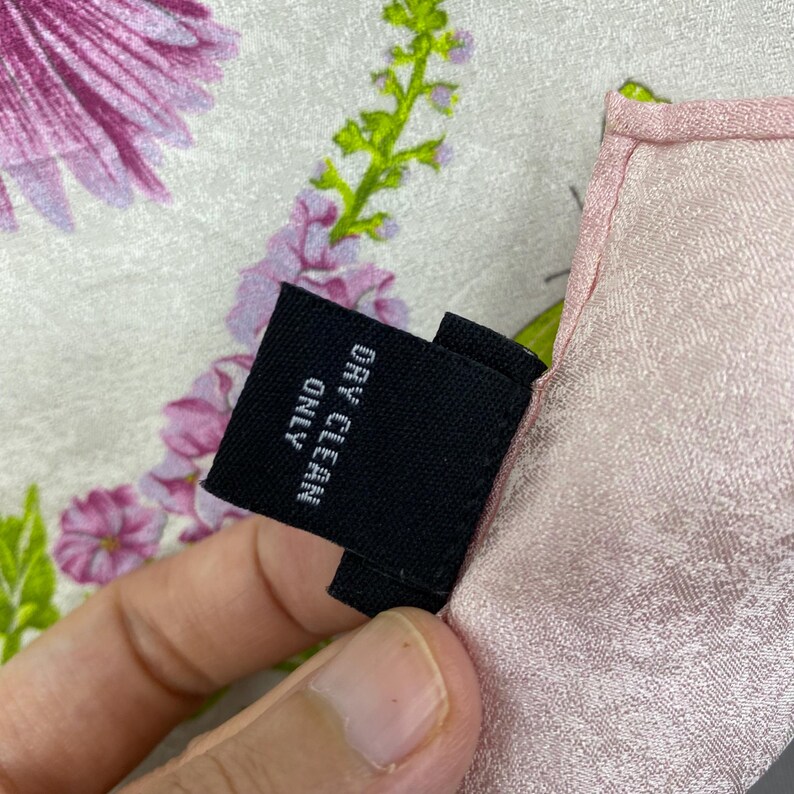 Puede incluir: Primer plano de una bufanda con estampado floral y borde rosa. La bufanda presenta un dise&ntilde;o de flores moradas y rosas con tallos verdes sobre un fondo blanco. Una etiqueta negra con las palabras "DRY CLEAN ONLY" es visible.