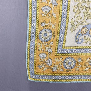 Puede incluir: Un textil cuadrado que presenta un panel central blanco con un patr&oacute;n floral de cachemira en azul claro y amarillo. El borde consiste en una banda azul claro con un motivo de flores de cuatro p&eacute;talos, enmarcado por una secci&oacute;n amarilla con enredaderas.