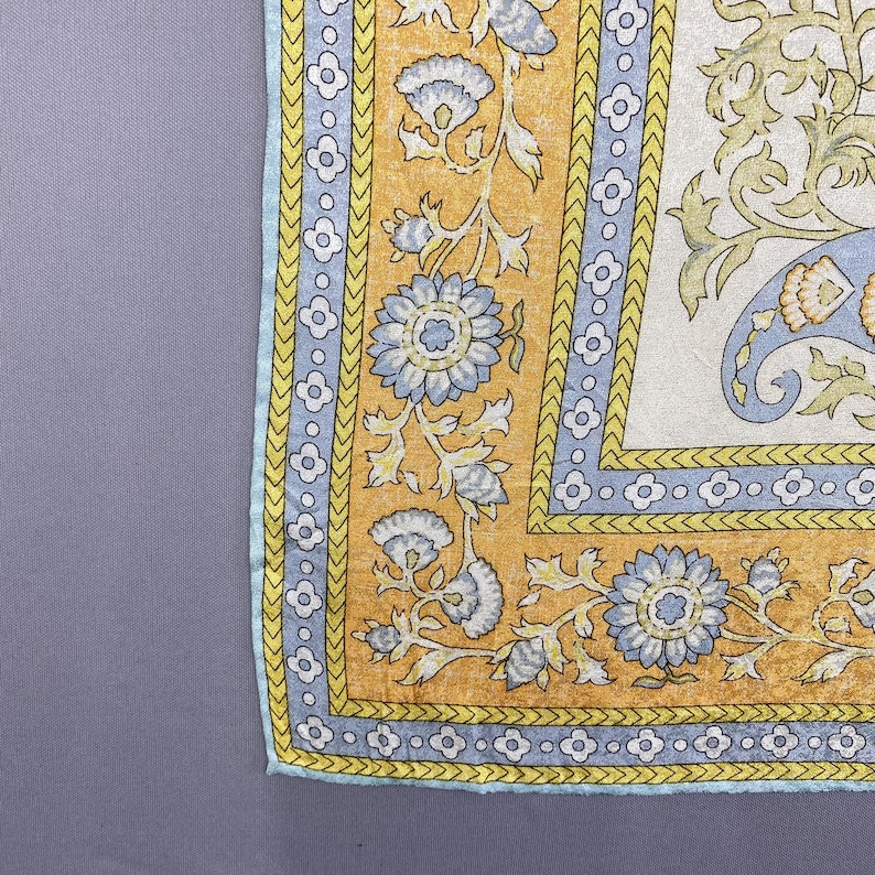 Puede incluir: Un textil cuadrado que presenta un panel central blanco con un patr&oacute;n floral de cachemira en azul claro y amarillo. El borde consiste en una banda azul claro con un motivo de flores de cuatro p&eacute;talos, enmarcado por una secci&oacute;n amarilla con enredaderas.