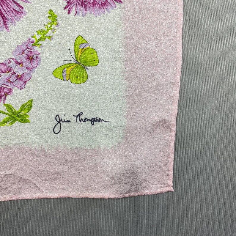 Puede incluir: Un pa&ntilde;uelo de seda rosa claro con un dise&ntilde;o floral y de mariposas. El pa&ntilde;uelo tiene un borde rosa m&aacute;s oscuro. El dise&ntilde;o incluye flores moradas, hojas verdes y una mariposa amarilla y verde. La firma "Jim Thompson" es visible.