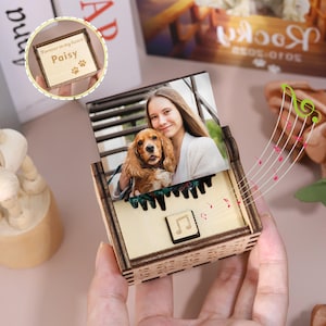Può includere: Un carillon in legno con una foto di una persona e di un cane. La scatola ha un design a tasti di pianoforte e un simbolo di nota musicale. La scatola è incisa con le parole "Forever in my heart" e il nome "Paisy" con impronte di zampe.