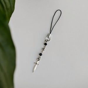 Może przedstawiać: Srebrny charms do telefonu z małą zawieszką w kształcie sztyletu. Charms zawiera czarne koraliki i czarną pętlę ze sznurka. Sztylet ma szczegółową rękojeść i spiczaste ostrze.