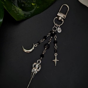 May include: A silver-toned metal keychain featuring a lobster clasp and three dangling chains. The chains are adorned with black beads, a silver crescent moon charm, a silver bird skull pendant, and a small four-pointed star charm.