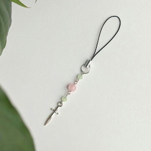May include: A silver-toned dagger charm with a black loop, pink and green beads. The charm is attached to a black cord, suitable for use as a phone charm or accessory. The background is white.
