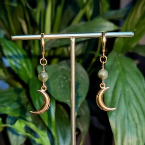 Puede incluir: Pendientes dorados con un dije de media luna y una cuenta redonda verde. Los pendientes cuelgan de un gancho dorado. El fondo es una imagen borrosa de follaje verde.