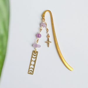 Puede incluir: Un marcapáginas de metal dorado con un diseño de fases lunares, una hebra colgante de cuentas moradas y un dije de estrella. El marcapáginas está diseñado para insertarse en un libro para marcar una página.