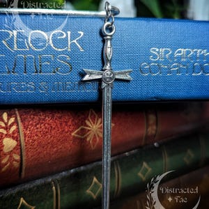 May include: A silver-toned metal sword pendant featuring a cross guard with a circular emblem at the centre. The sword has a straight, double-edged blade and a detailed hilt. A small metal jump ring is attached to the top of the sword pommel.
