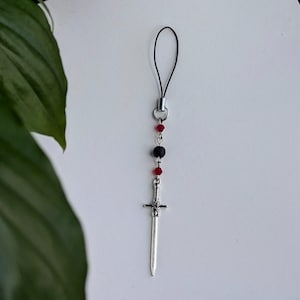 May include: A phone charm featuring a silver-toned sword pendant suspended from a black cord. The design includes two faceted red beads and one matte black bead arranged vertically on a silver metal chain, creating a linear decorative accessory.