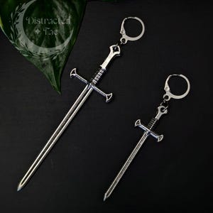 May include: Two silver-toned sword-shaped earrings with lever-back closures. The earrings feature a detailed hilt and a long, straight blade design. One sword is noticeably longer than the other, and both have a metallic finish with dark accents.