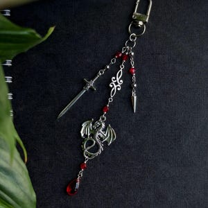 May include: Silver-toned keychain featuring a sword, dragon, and decorative elements. Red beads and a teardrop-shaped charm add color. The keychain is designed with a lobster clasp for easy attachment.
