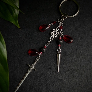 Peut inclure: Un porte-clés argenté avec une épée décorative, une pointe conique et des perles rouges en forme de goutte. Le porte-clés comporte également des perles noires et rouges et un motif argenté décoratif.