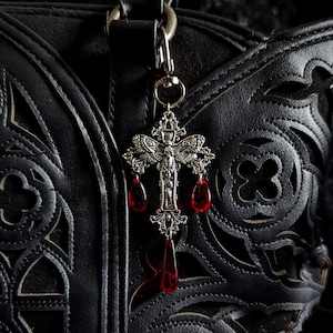 May include: A silver cross-shaped charm with red teardrop accents hangs from a black leather bag. The charm has detailed patterns and is attached to a silver clasp. The bag features a gothic design with cut-out patterns.