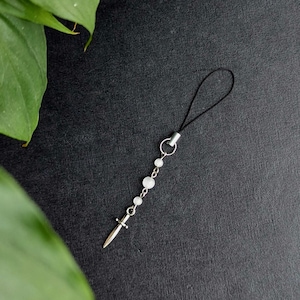 May include: A phone charm featuring a silver-toned metal sword pendant hanging from a delicate chain with three small, round, translucent white beads. The charm is attached to a thin black cord loop for easy attachment to a mobile device.