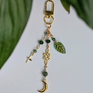 May include: Gold-colored keychain with a lobster clasp. The charm features a crescent moon, a star, a leaf, and decorative gold filigree. Green beads and accents add color to the design. The background is a soft white with green foliage.