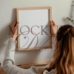 May include: A rectangular wooden picture frame with a white interior is held by a person. The frame is light brown and the text "MOCK Deluxe" is printed on the white surface. The person is wearing a light gray sweater.