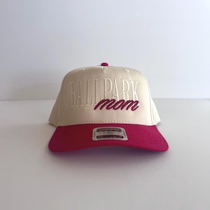 May include: A baseball cap with a cream-colored crown and a magenta brim. The text "BALLPARK mom" is embroidered on the front in magenta. The cap has a curved brim and a small button on top.