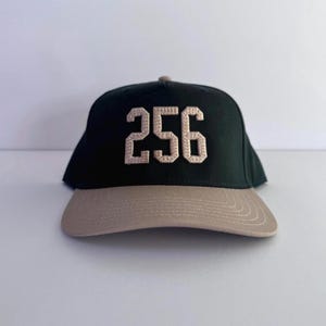 May include: A dark green baseball cap with a tan brim. The number "256" is embroidered in a tan color on the front of the cap. The cap is set against a white background.