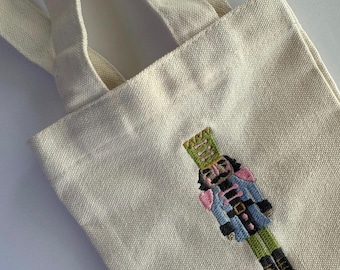 May include: Cream-colored canvas tote bag with embroidered nutcracker design. The nutcracker is depicted in blue, pink, green, and black. The bag has two cream-colored handles.