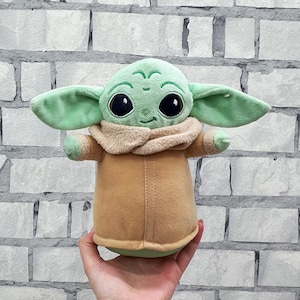 May include: A plush toy of a character with large eyes, green skin, and large ears. The character is wearing a tan coat and a beige collar. The toy is being held in a person's hand against a white brick wall.