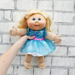 Könnte beinhalten: Eine Vintage-Puppe mit blonden Haaren, blauen Augen und einem lächelnden Gesichtsausdruck. Die Puppe trägt ein blaues Kleid mit einem rosa Gürtel und einem hellblauen Rock. Die Puppe wird vor einer weißen Backsteinmauer gehalten.