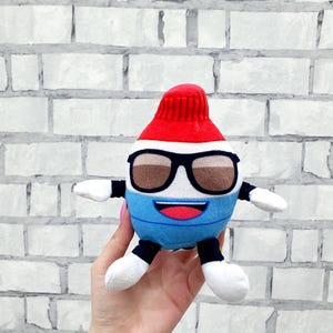 May include: A plush toy shaped like a tube of toothpaste, featuring a red cap, black-framed sunglasses, and a blue and white body. The toy has white arms and legs, and a smiling mouth.