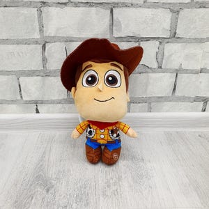 May include: A soft toy of the character Woody from Toy Story. The toy features a tan face, brown hair and hat, and is dressed in a yellow plaid shirt, blue trousers, and brown boots. It stands in front of a white brick wall.