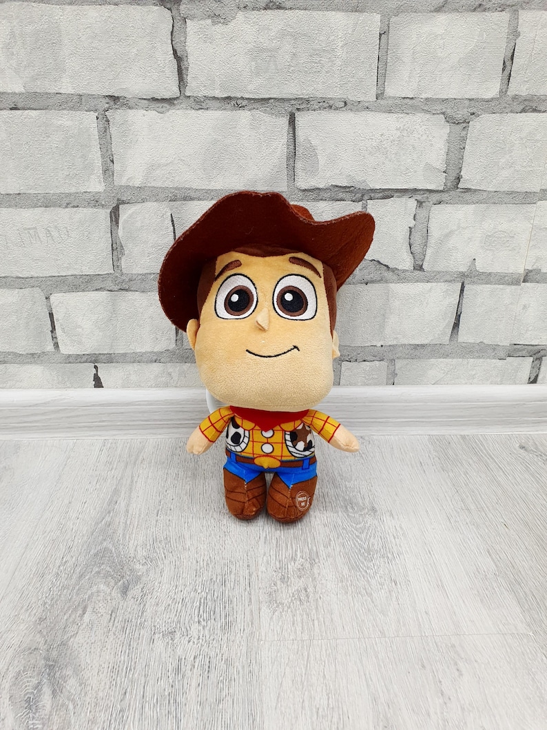 May include: A soft toy of the character Woody from Toy Story. The toy features a tan face, brown hair and hat, and is dressed in a yellow plaid shirt, blue trousers, and brown boots. It stands in front of a white brick wall.