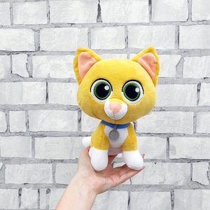May include: A plush yellow cat with large green eyes, pink inner ears, and a white muzzle. It has a blue collar with a silver tag. The cat's paws and the tip of its tail are white. The background is a white brick wall.