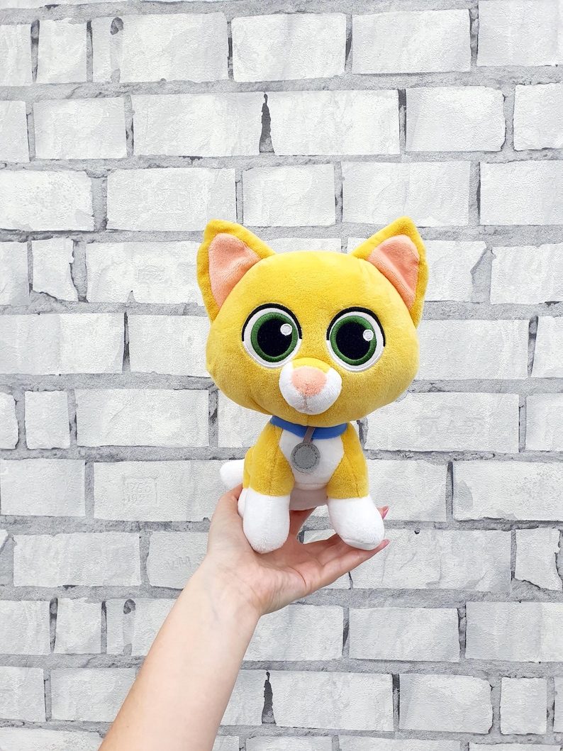 May include: A plush yellow cat with large green eyes, pink inner ears, and a white muzzle. It has a blue collar with a silver tag. The cat's paws and the tip of its tail are white. The background is a white brick wall.
