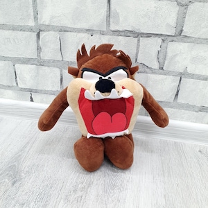 May include: A plush toy of the Tasmanian Devil, a cartoon character. The toy is brown with a tan face, large red mouth, and black nose. It has small arms and legs, and is posed in a standing position.