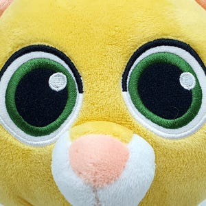 May include: Close-up of a plush toy head with large, expressive eyes. The toy features bright green eyes with white highlights, outlined in black and white stitching. The plush is primarily yellow with peach-colored ears and a pink nose.