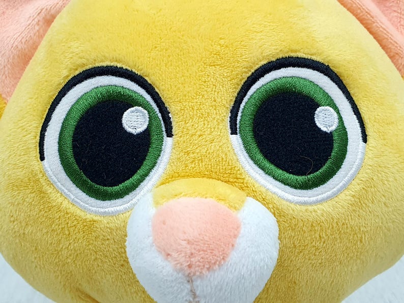 May include: Close-up of a plush toy head with large, expressive eyes. The toy features bright green eyes with white highlights, outlined in black and white stitching. The plush is primarily yellow with peach-colored ears and a pink nose.