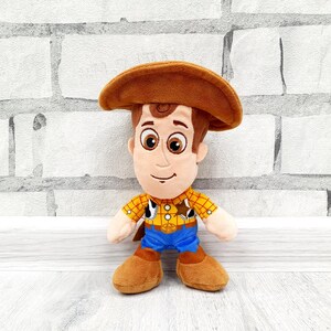 K&ouml;nnte beinhalten: Ein Pl&uuml;schtier einer Cartoon-Cowboy-Figur mit braunem Cowboyhut, gelbem kariertem Hemd, blauer Hose und braunen Stiefeln. Das Spielzeug hat einen freundlichen Gesichtsausdruck und steht aufrecht.